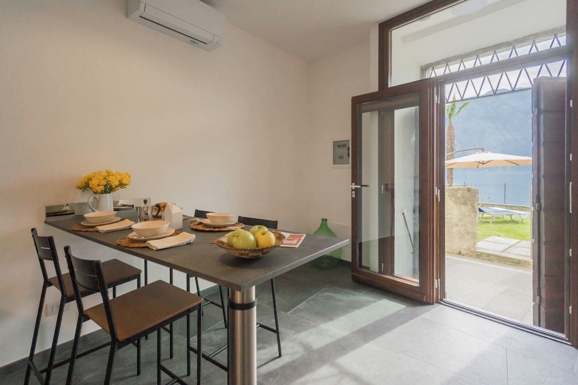Un comedor con una mesa dispuesta para dos personas, un frutero y un jarrón con flores amarillas.