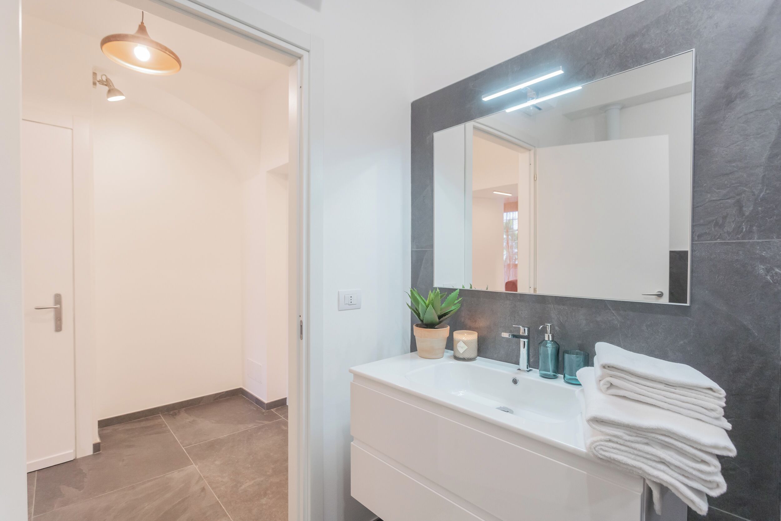 A modern bathroom with a white vanity, a large mirror, a potted plant, and a stack of towels.