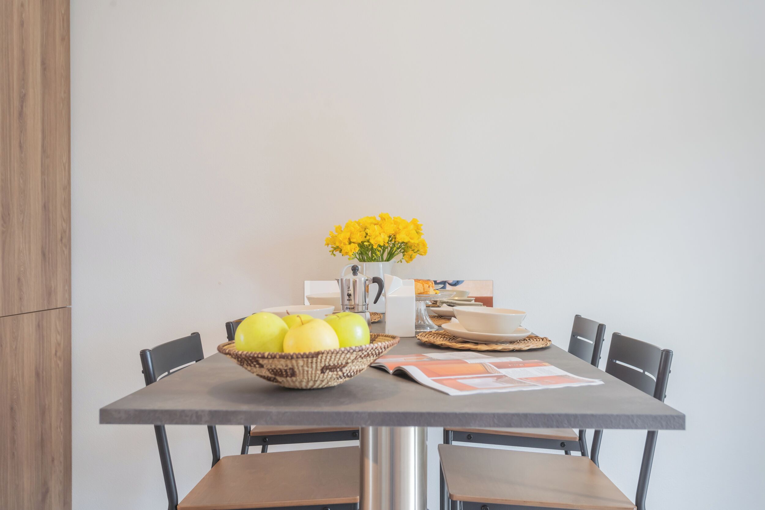 A dining table with a basket of yellow apples, a vase of yellow flowers, and a newspaper.