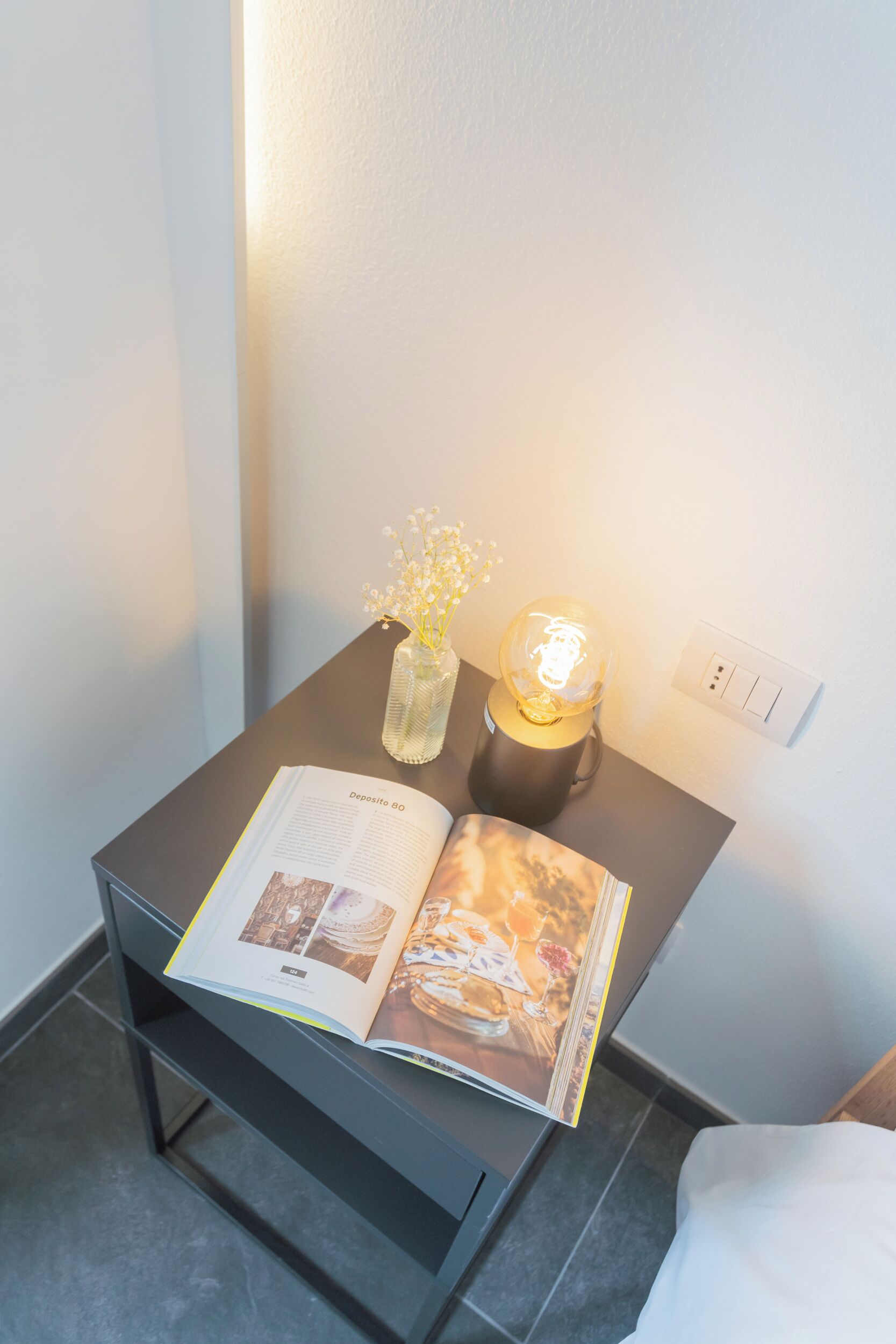 A neatly arranged bedside table with a book, a vase with flowers, and a lamp.