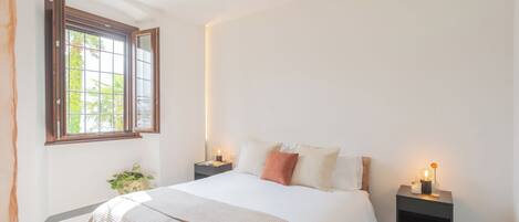 A bedroom with a large bed, two bedside tables, a window with a view, and a small plant.