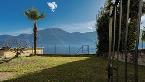 A grassy area with a palm tree, a metal fence, and mountains in the background. - Family Lake Apartment by Rent all Como (Tremezzina)