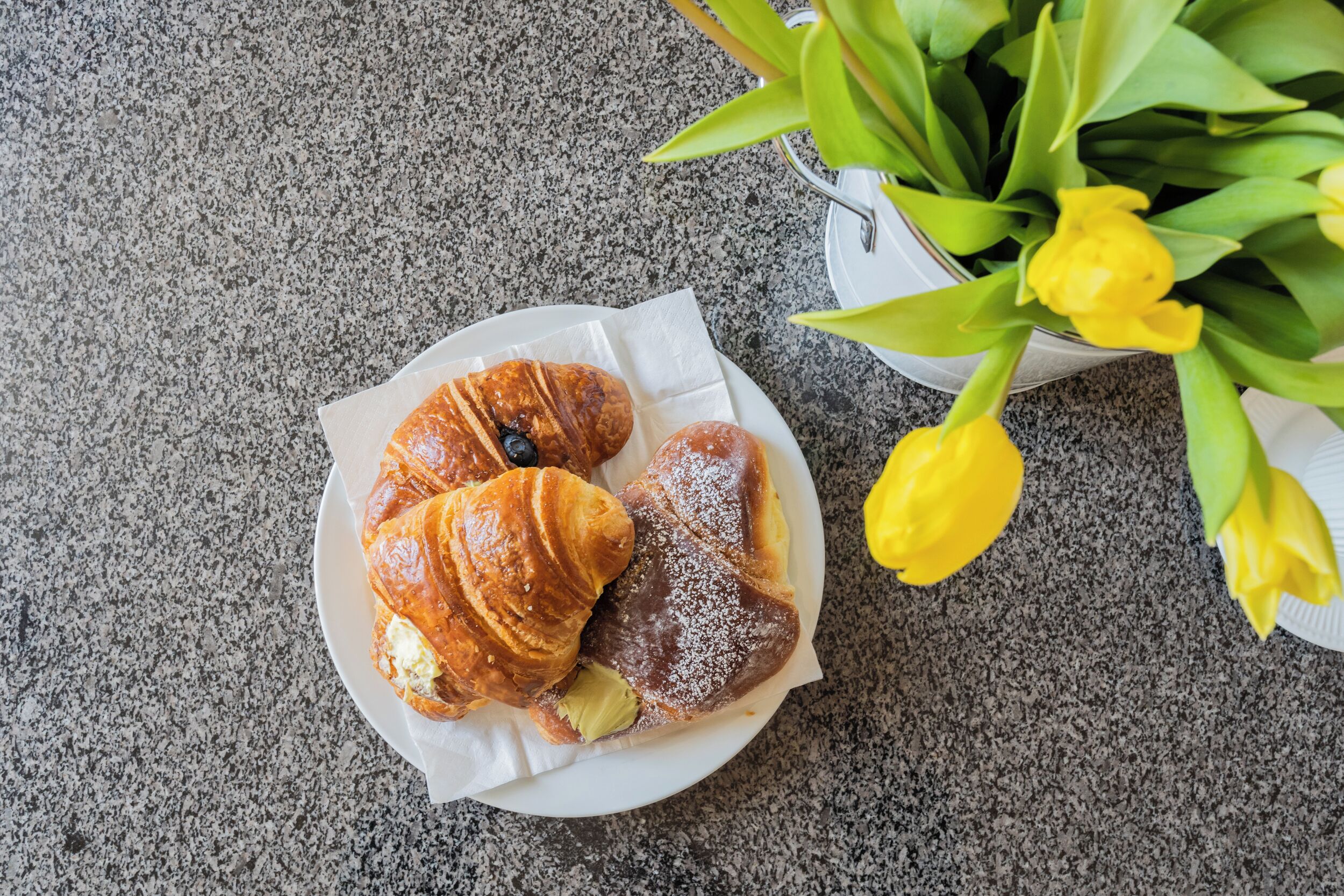 A plate with croissants and a pastry on a granite countertop, accompanied by a potted plant with yellow flowers.