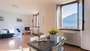 A modern living room with a blue sofa, a dining table with chairs, a TV, and a large window with a mountain view. - Family Lake Apartment by Rent all Como (Tremezzina)