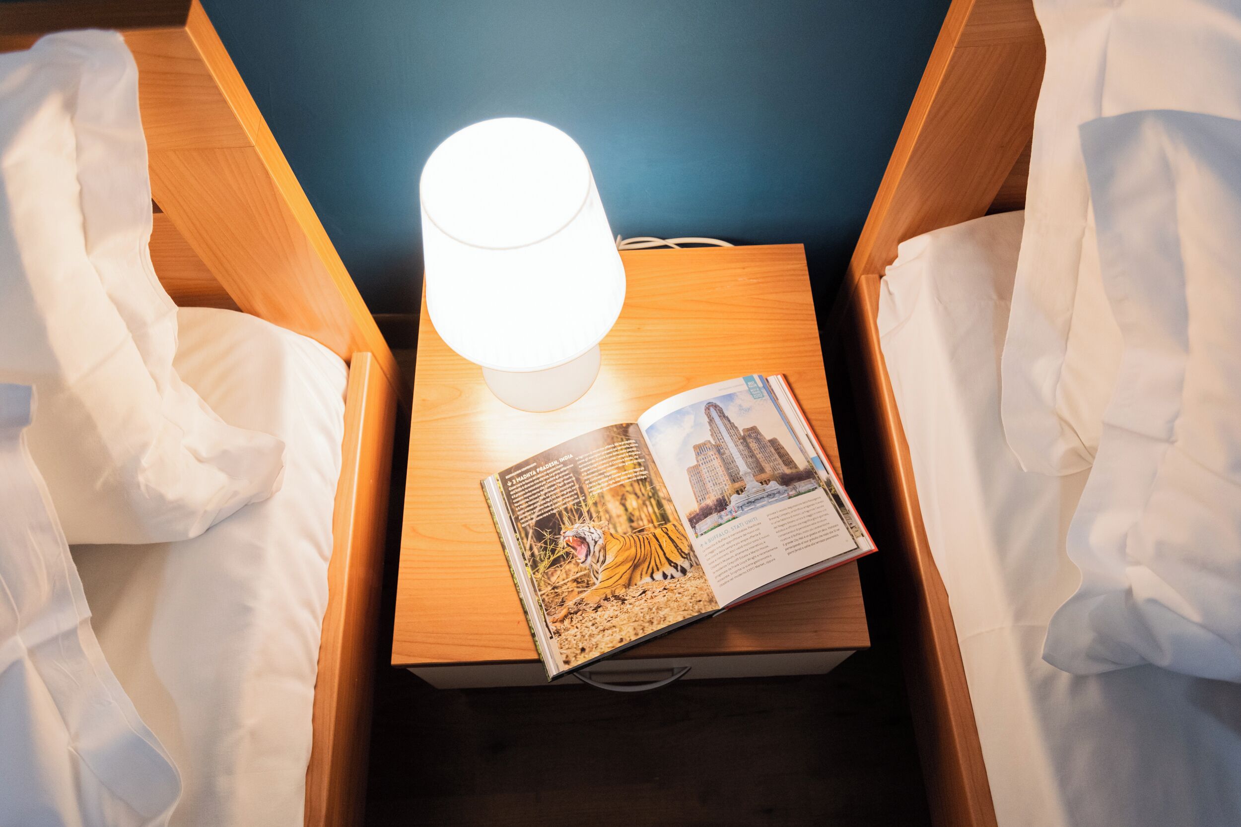 Une chambre d’hôtel avec une table de chevet en bois, une lampe blanche et un livre dont la couverture représente un paysage urbain.
