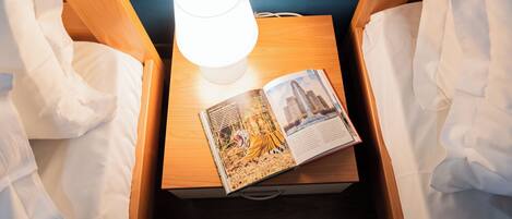 Une chambre d’hôtel avec une table de chevet en bois, une lampe blanche et un livre dont la couverture représente un paysage urbain.