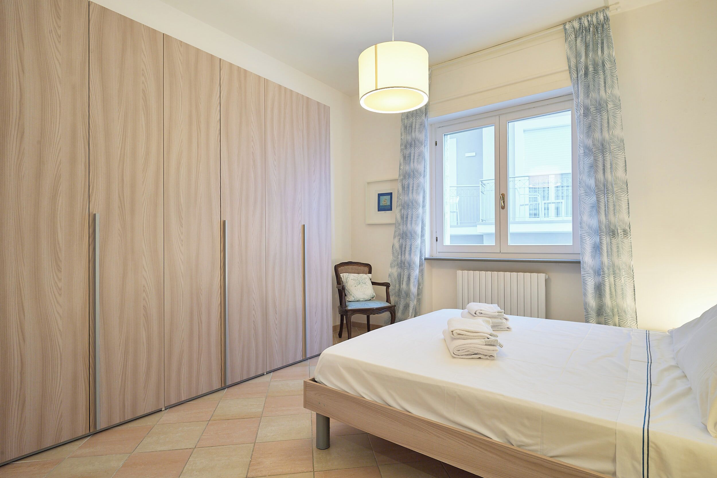 A bedroom with a bed, a chair, a window with curtains, and a wardrobe.