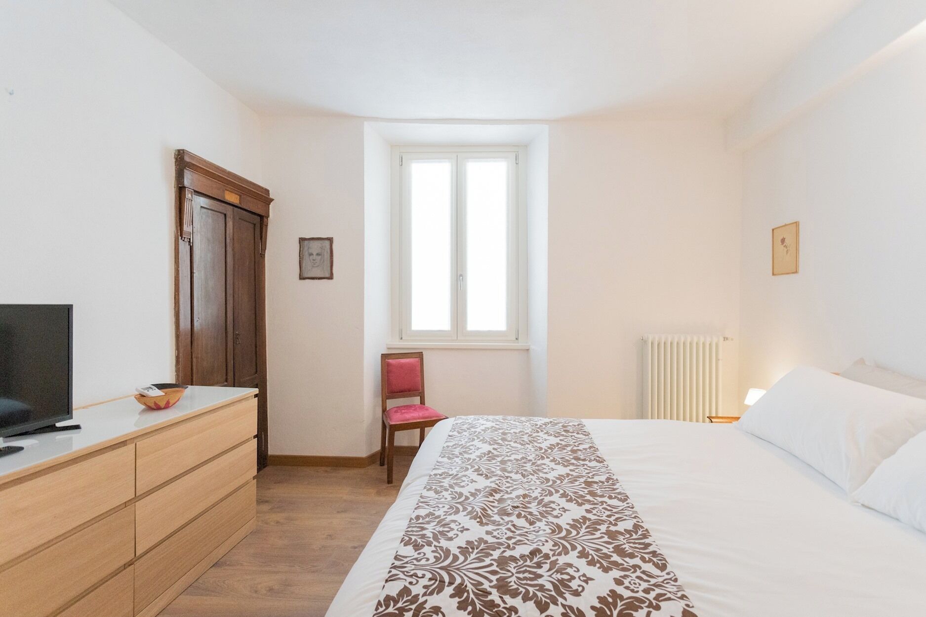 Une chambre à coucher avec un lit, une commode, une chaise, une fenêtre et un radiateur.