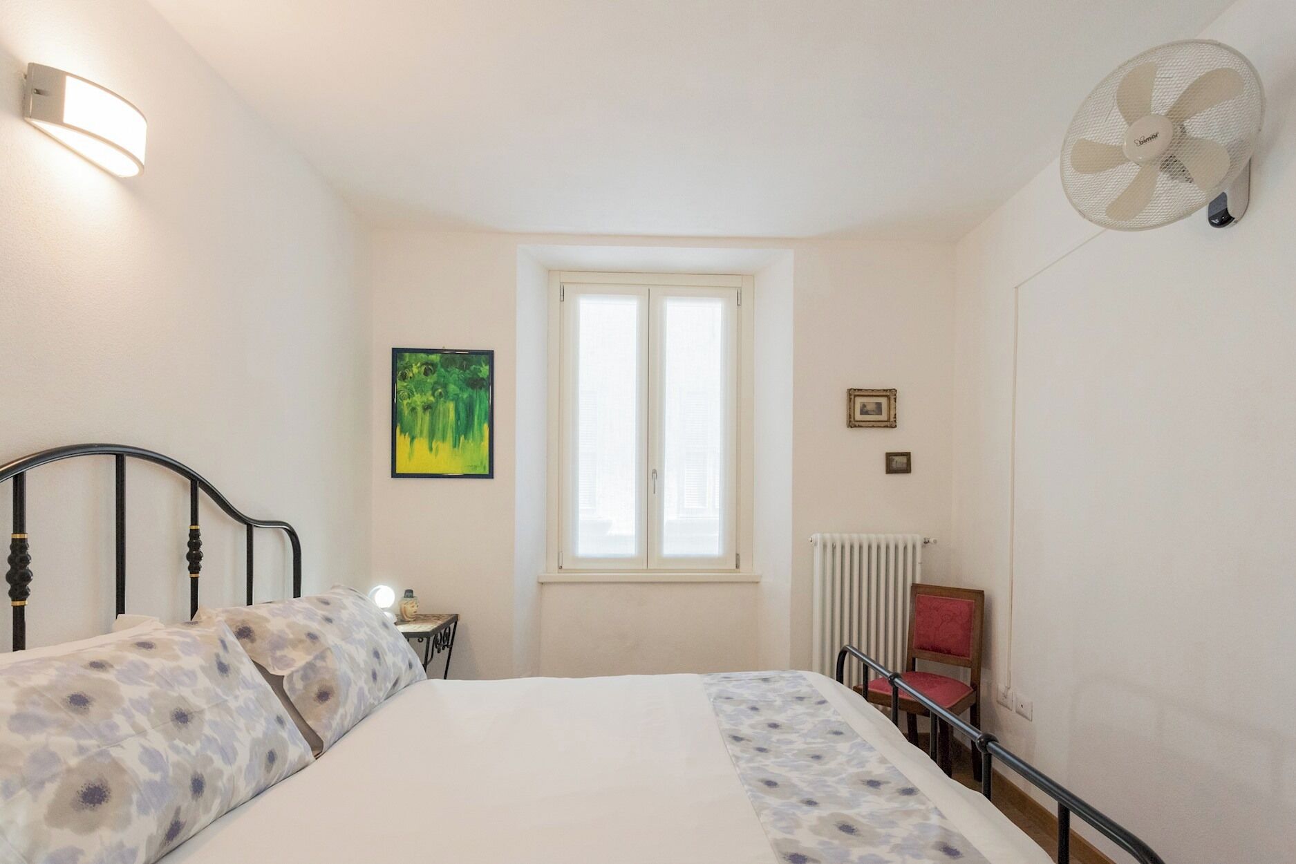 Una camera da letto con un letto, un comodino, una finestra, un termosifone e una ventola.