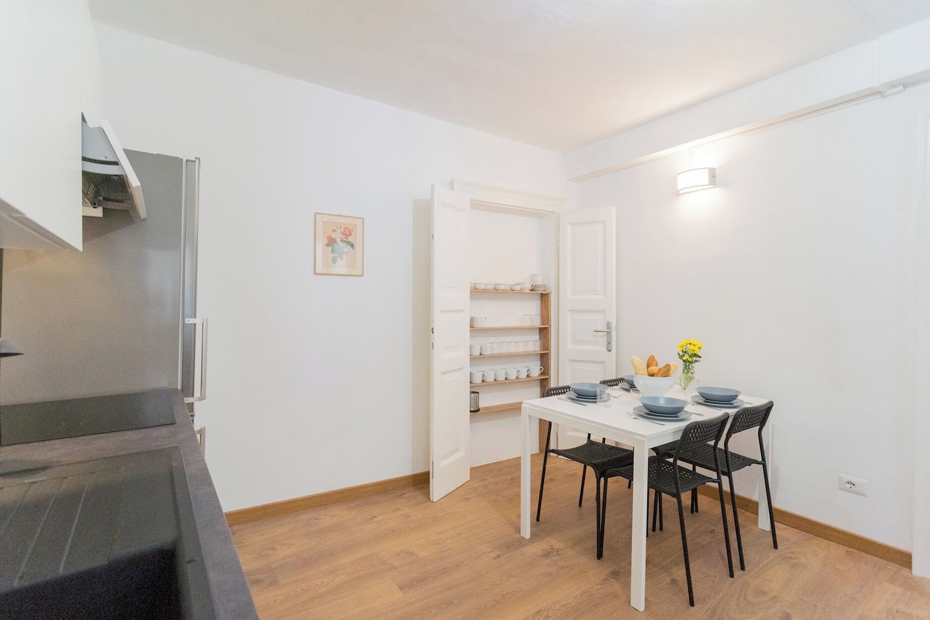 A compact kitchen with a built-in oven, a dining area with a white table and black chairs, and a white door leading to a storage space with shelves.