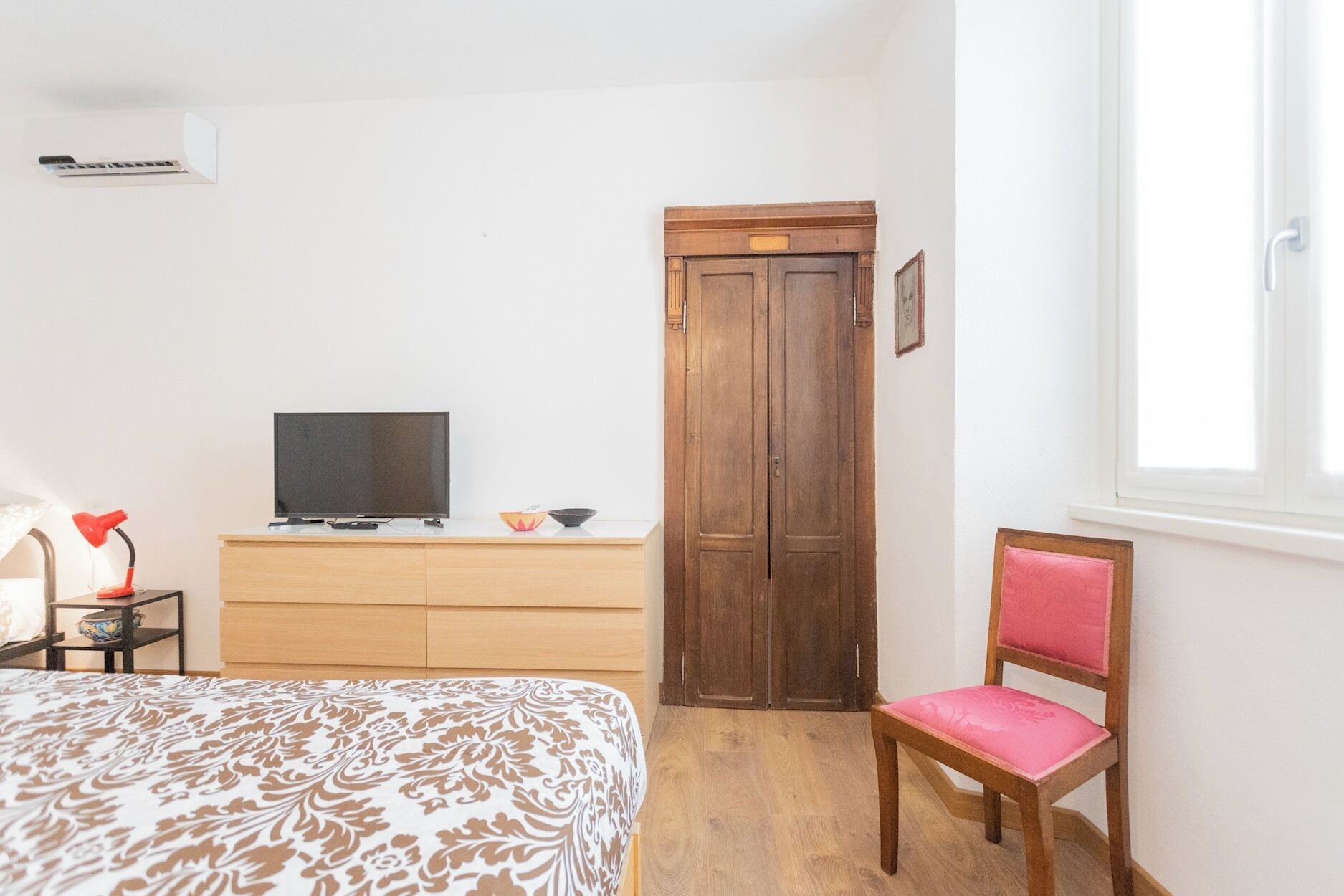 Ein Schlafzimmer mit einem Bett, einem Fernseher auf einer Holzkommode, einem Schrank aus Holz, einem Sessel und einem Fenster.