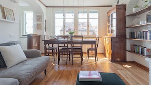 Una sala de estar acogedora con suelo de madera, un sofá gris, una mesa de comedor con sillas, una estantería y una ventana con vistas a edificios.