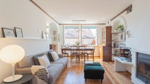 Una sala de estar acogedora con sofá, puf y una mesa de comedor con sillas. Hay una chimenea y una estantería.