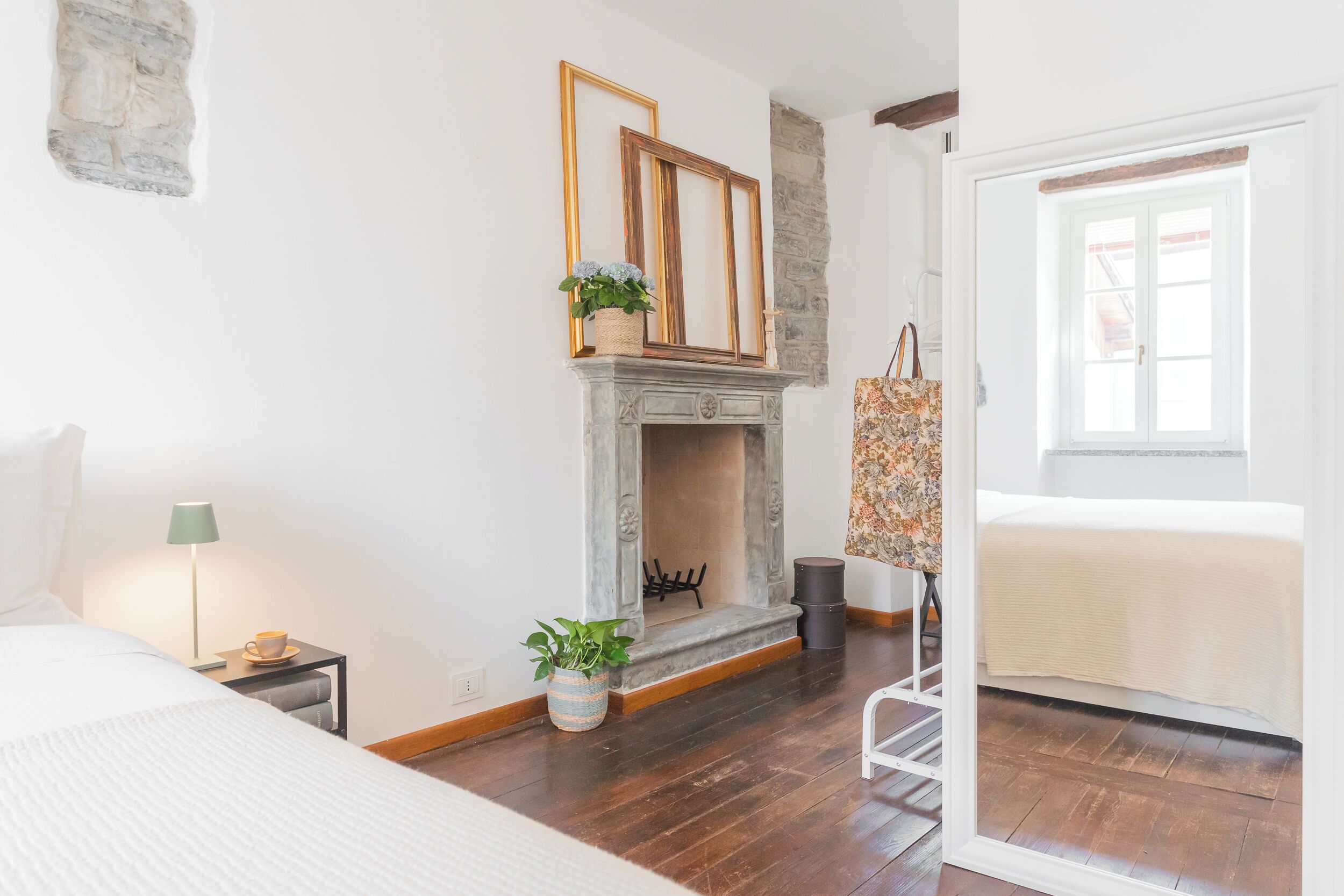 Un dormitorio con cama, mesita de noche con lámpara, chimenea, planta en maceta y ventana.
