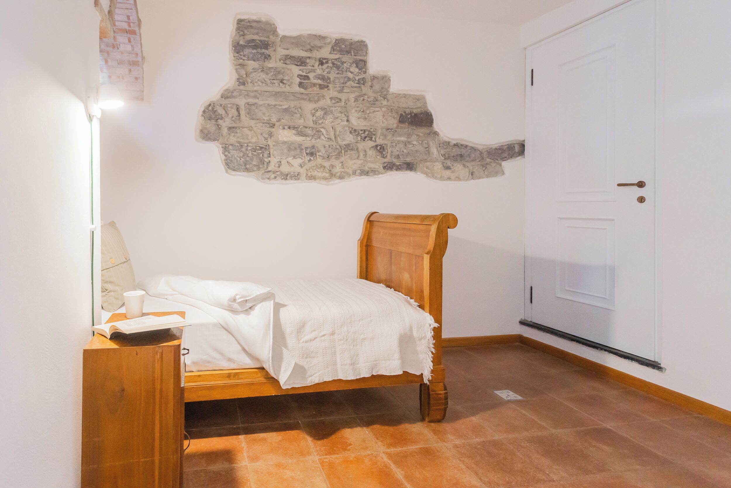 Quarto com cama de madeira, parede branca, parede de pedra e porta branca.