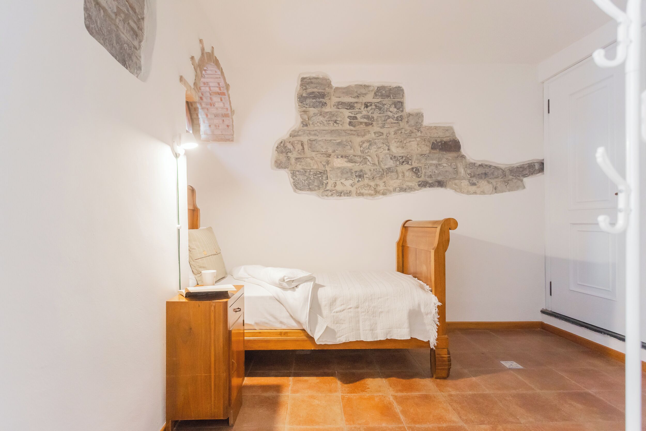A small, cozy bedroom with a single bed, a nightstand, a lamp, and a stone wall feature.