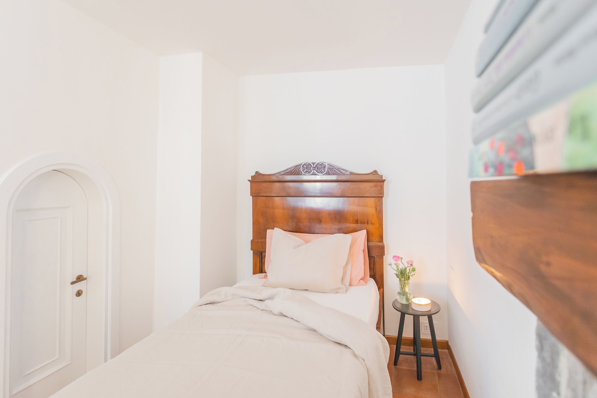 A narrow room with a wooden bed, a white door, and a small table with a candle.