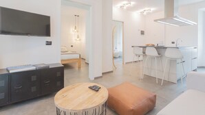 A modern living room with a TV, a wooden coffee table, and a sofa. There is a kitchen area with white bar stools and a large window. - Enchanted Como Lake - City Apartment by Rent all Como (Como)