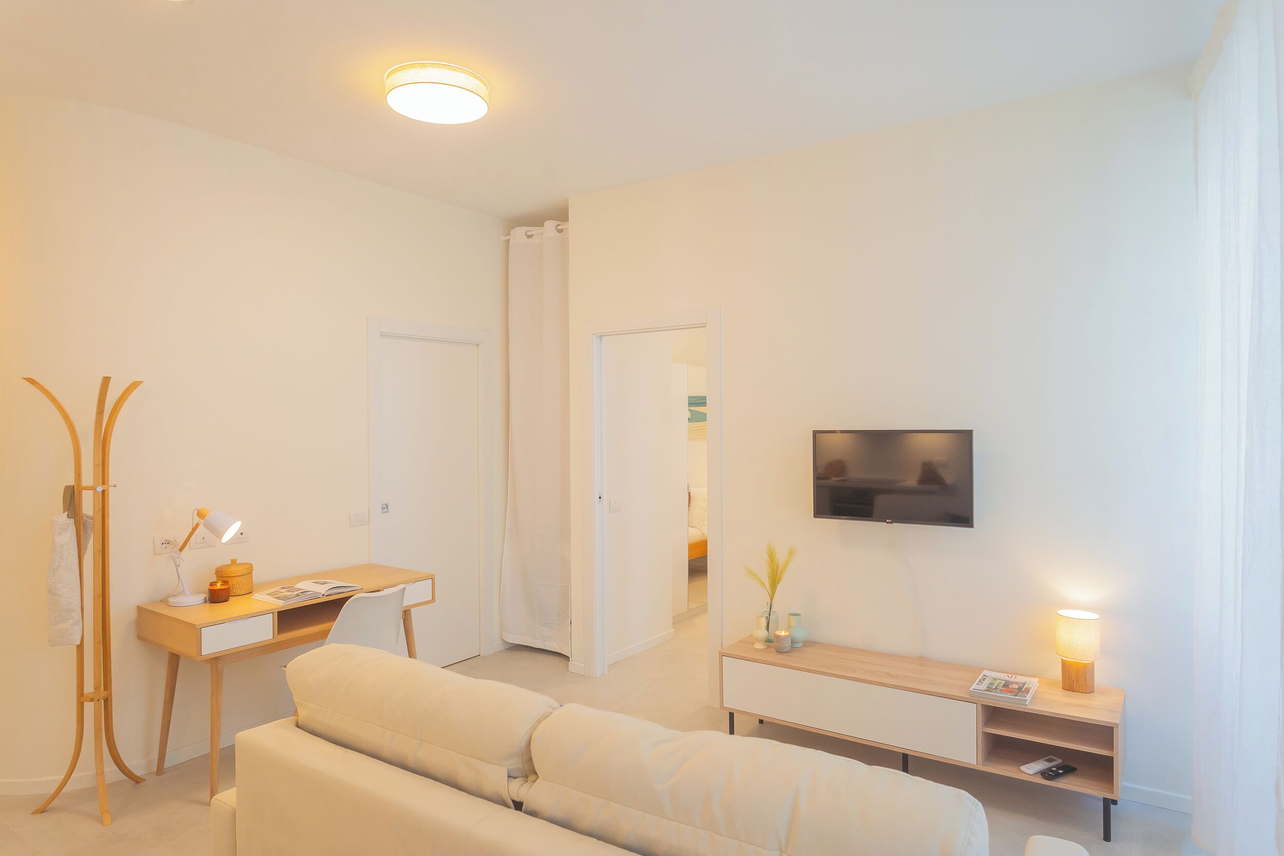 Una habitación de hotel moderna con sofá, escritorio con lámpara, televisor montado en la pared y una mesa consola de madera.