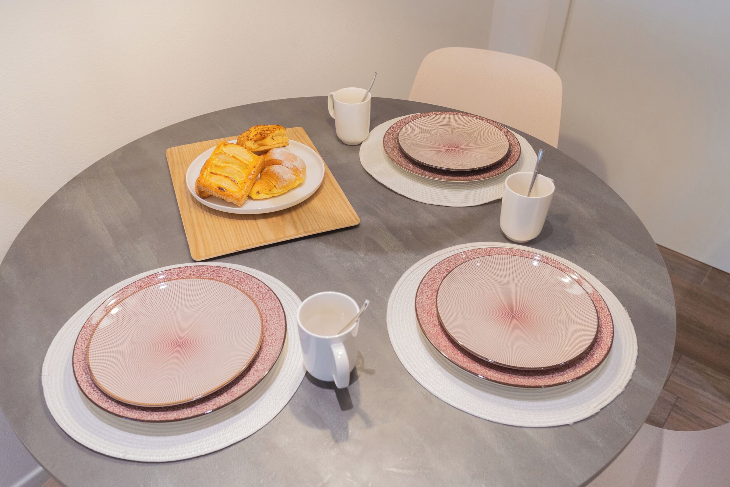 Uma mesa de jantar redonda com pratos rosa e brancos, uma tábua de madeira com doces e duas xícaras brancas.