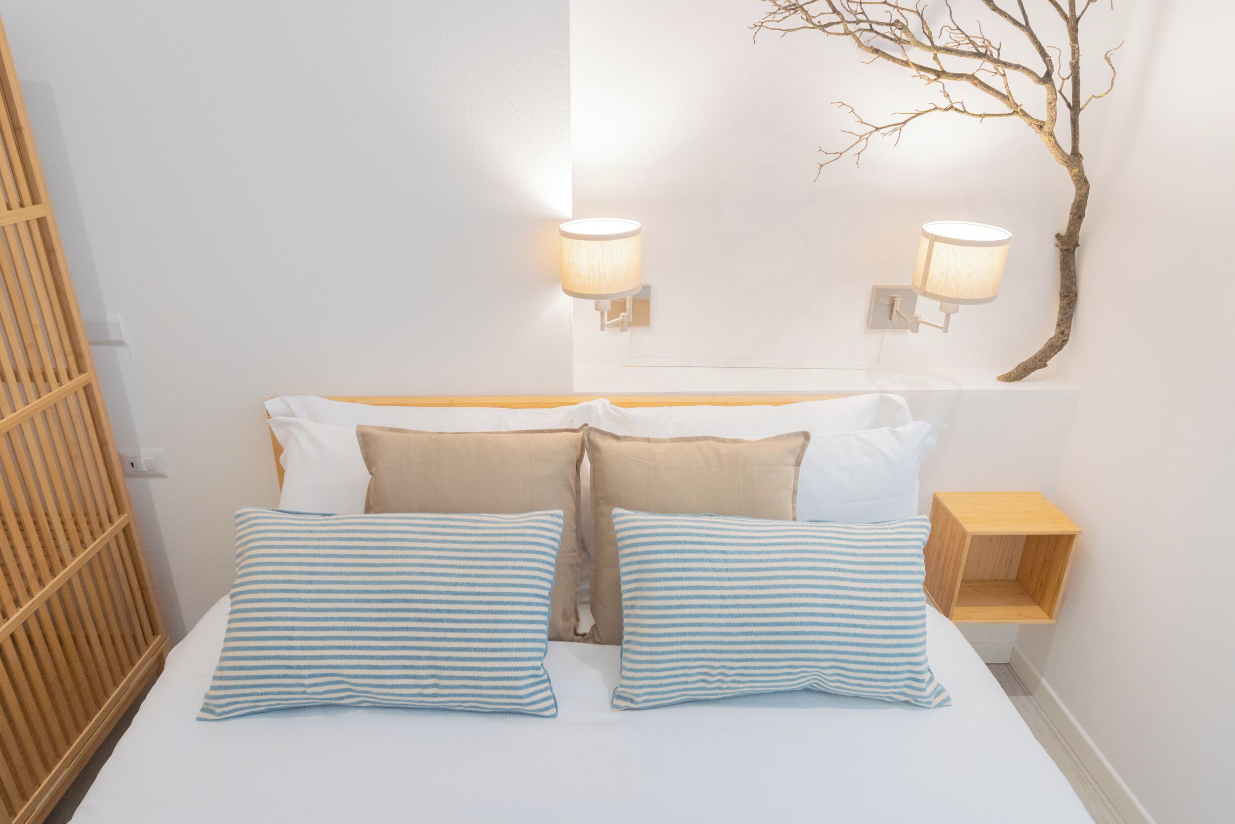 Een net opgemaakt bed met gestreepte kussens, een houten nachtkastje en wandlampen.