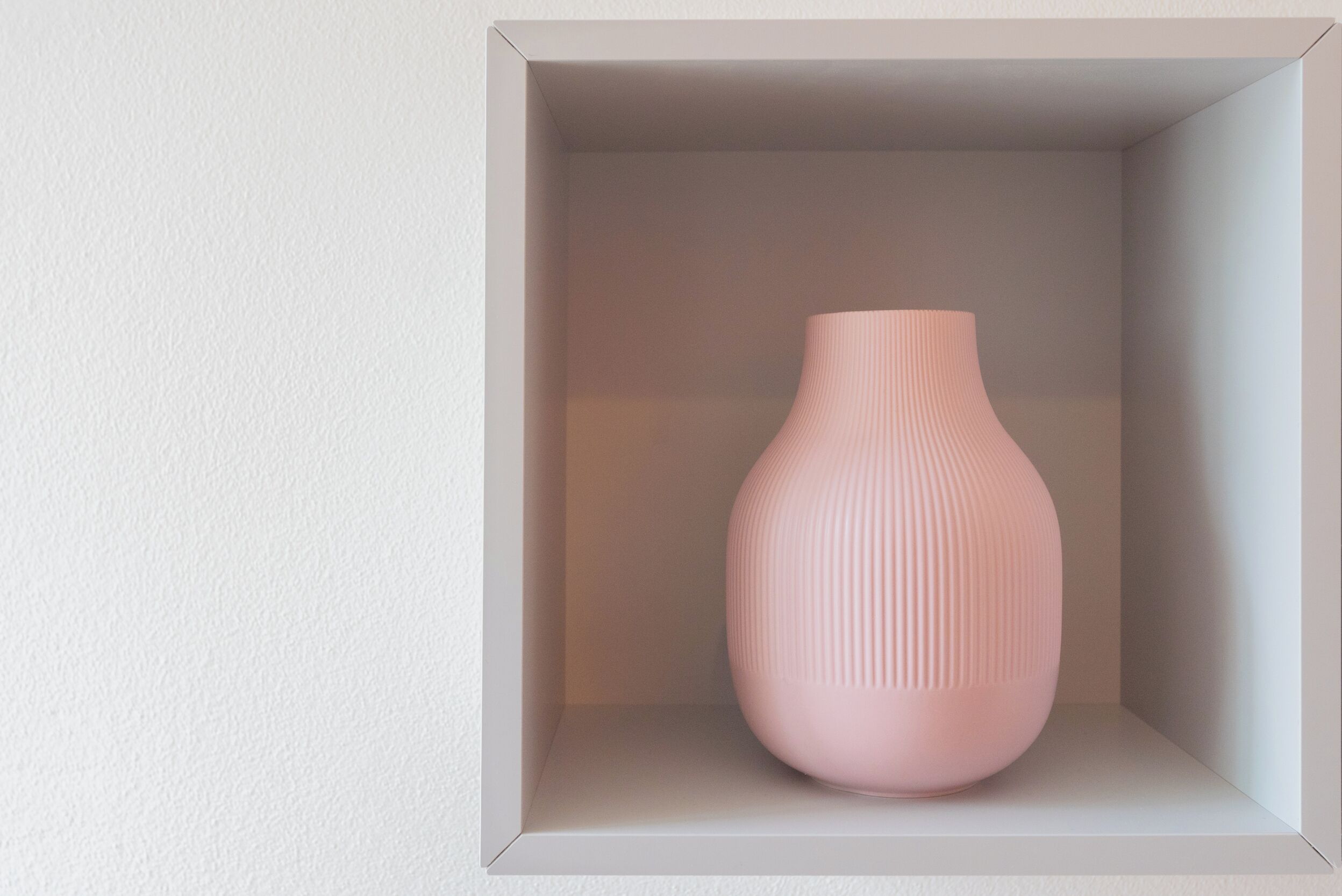 Un expositor blanco y cuadrado, vacío, con un jarrón rosa en su interior.
