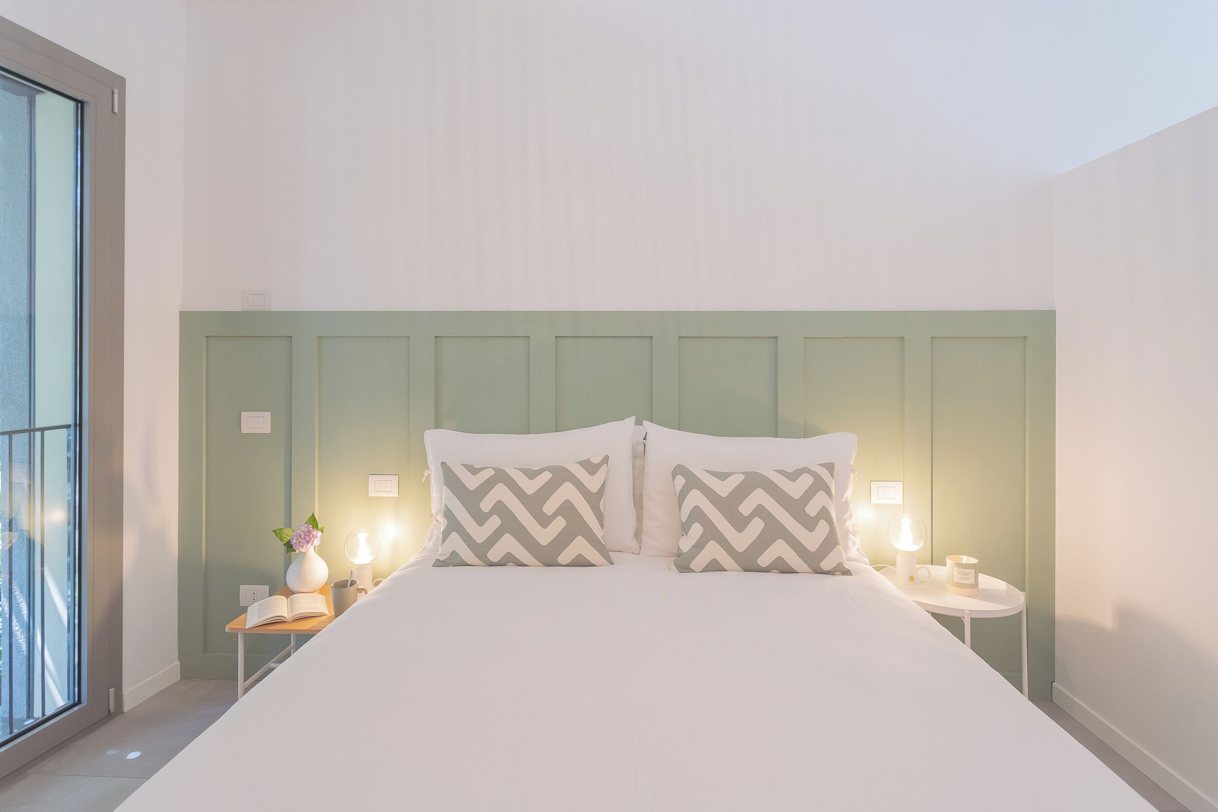 A neatly made bed with two pillows, a bedside table with a lamp, and a vase with flowers.