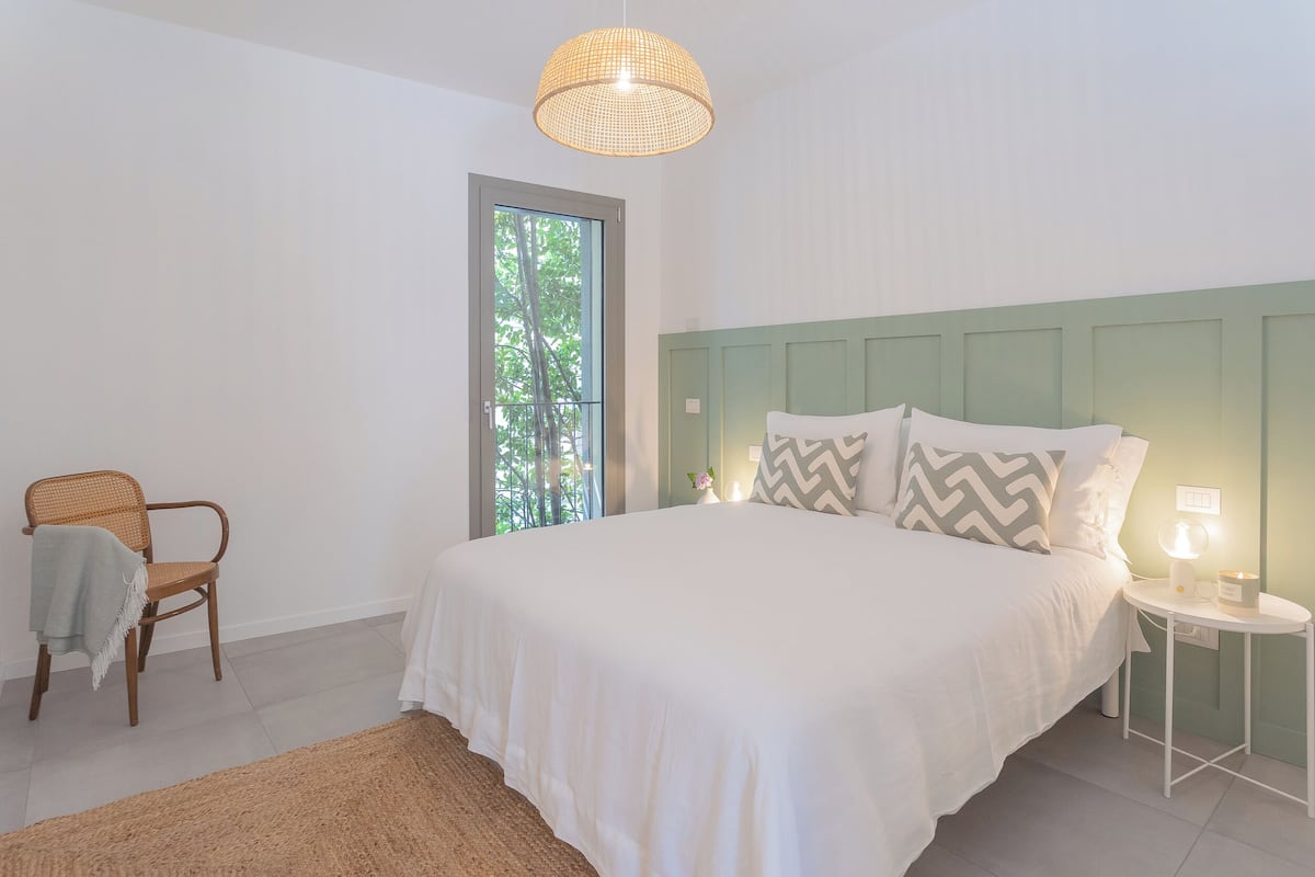 A bedroom with a large bed, a chair, a bedside table with a lamp, and a window with a view of trees.