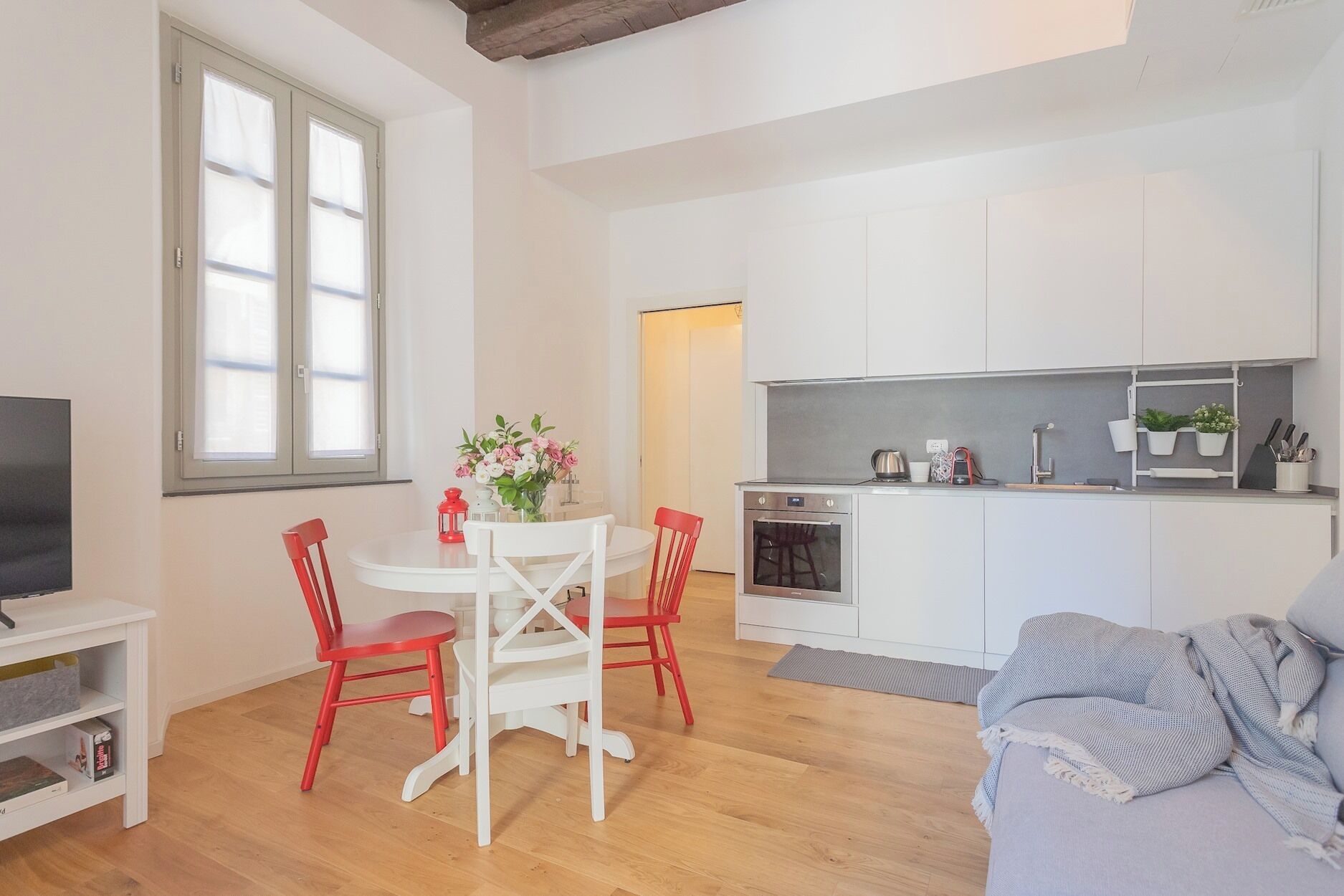 Una cucina moderna con mobili bianchi, forno integrato e un piccolo angolo ristoro con tavolo bianco e sedie rosse.