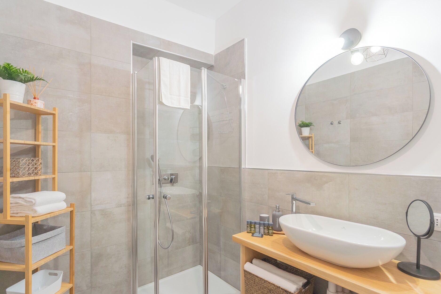 A modern bathroom with a glass-enclosed shower, a round mirror, and a wooden shelf with towels.
