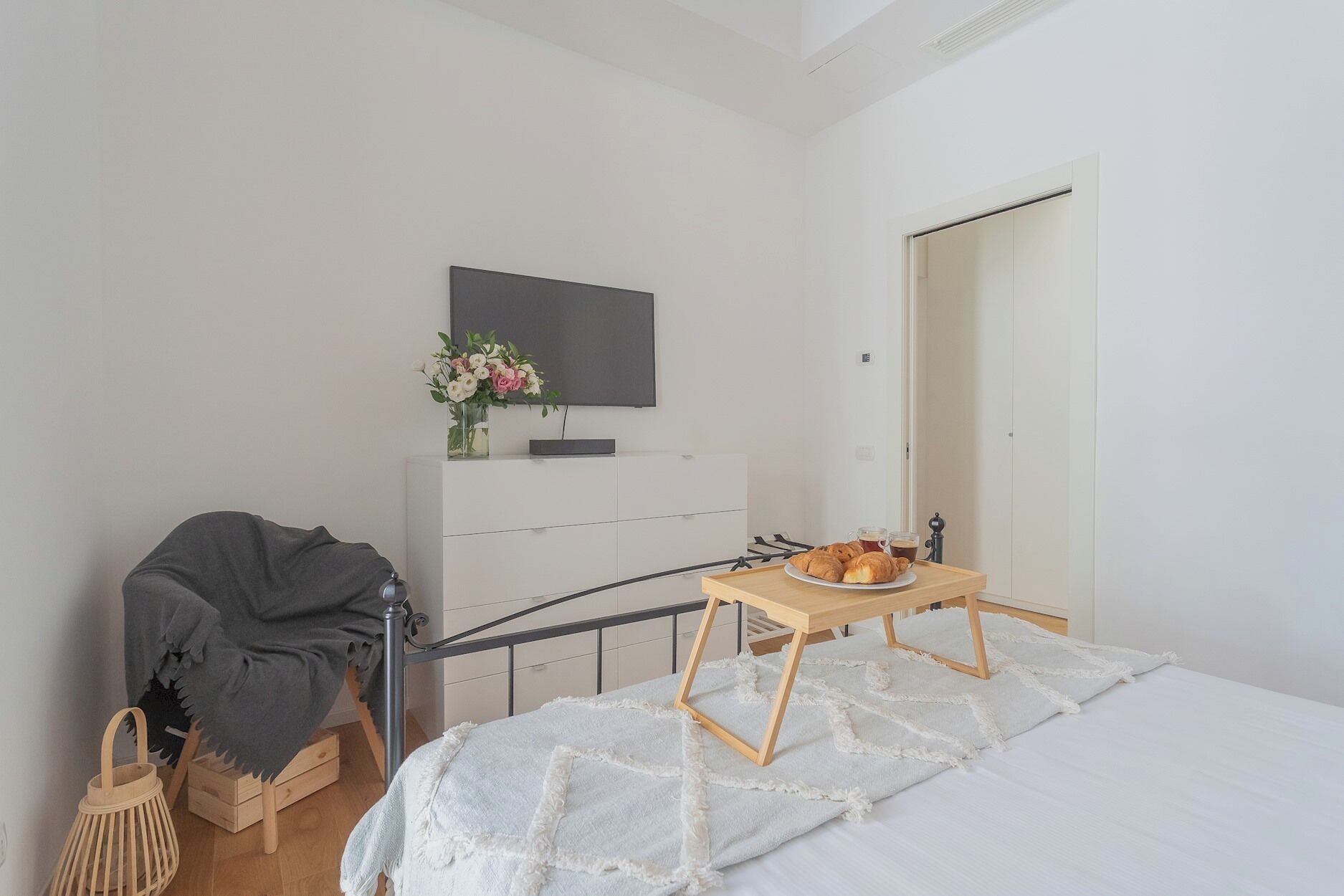 A bedroom with a bed, a bedside table with a tray of pastries, a TV mounted on the wall, and a wooden cabinet.