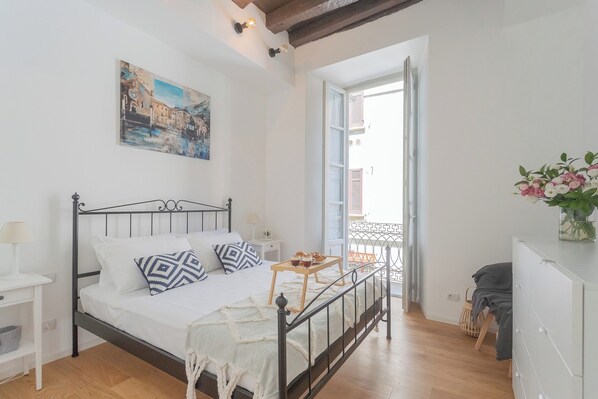 A bedroom with a bed, bedside tables, a painting, a balcony with a railing, and a view of buildings outside.