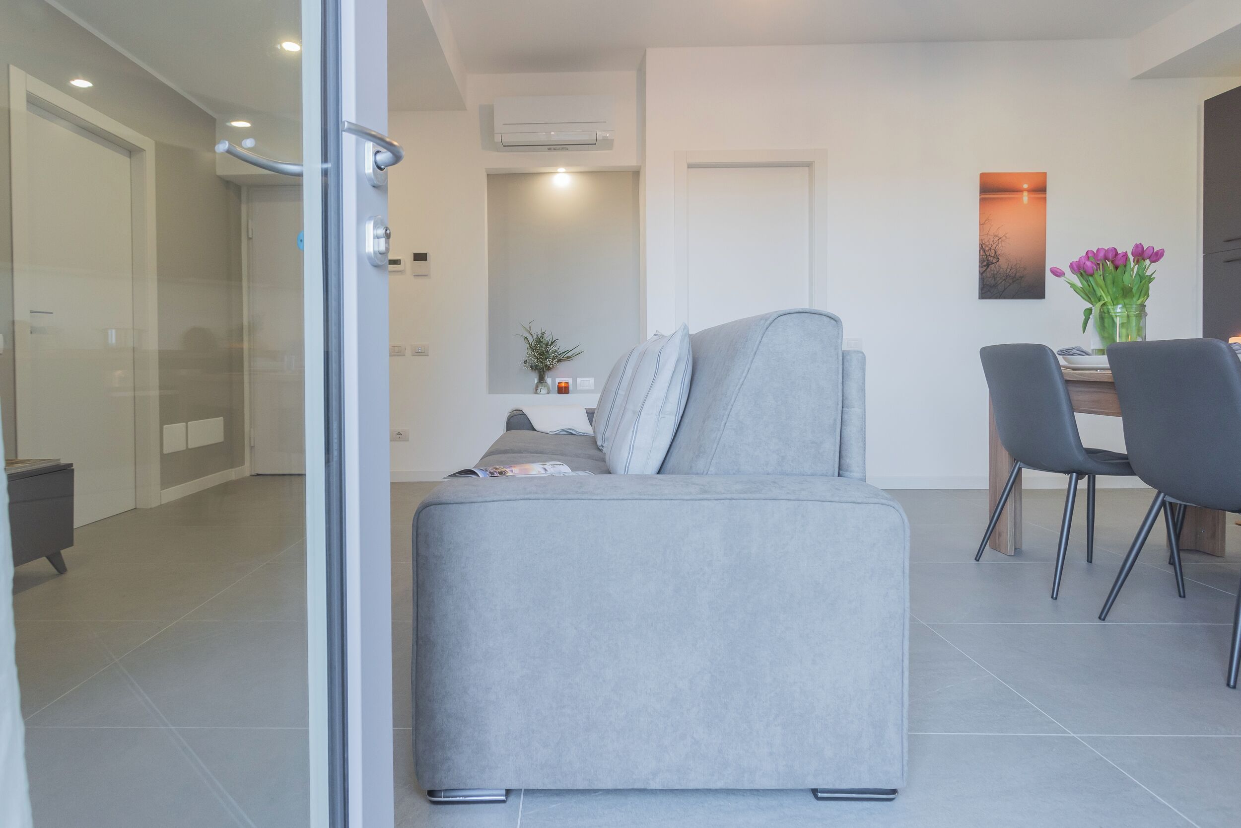 A modern living room with a grey sofa, a dining table with chairs, and a vase of flowers on the wall.