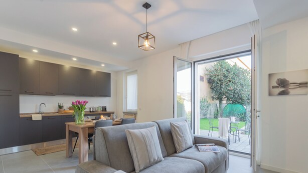 A modern living room with a grey sofa, a wooden dining table, and a kitchen in the background. - Isola Felice Grandate by Rent all Como (Grandate)