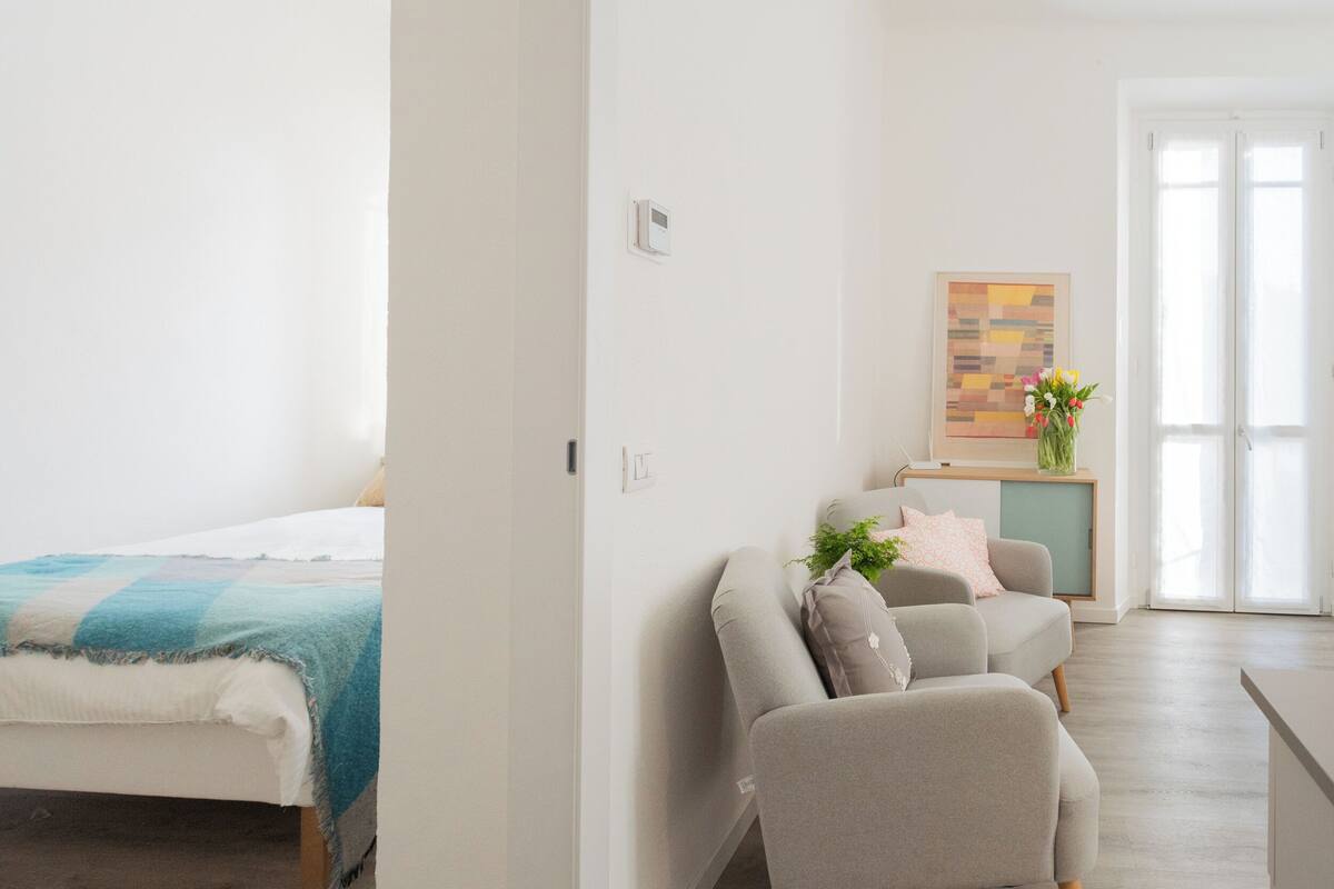A modern, well-lit room with a bed, a sofa, and a small kitchenette area.