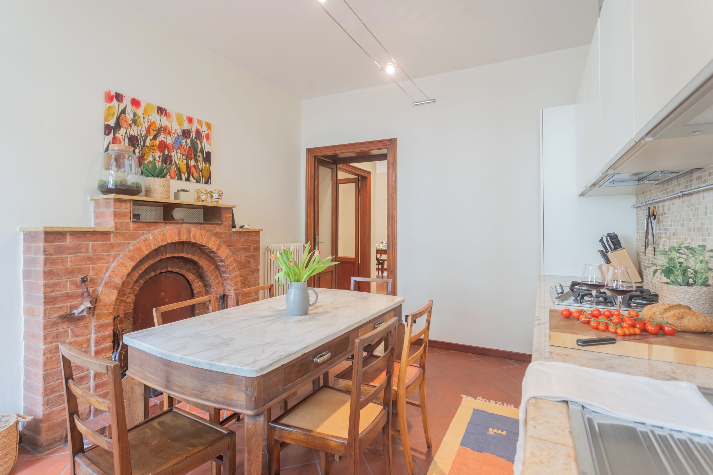 A kitchen with a brick fireplace, wooden dining table, and chairs. There is a countertop with a toaster, a knife block, and a basket of tomatoes.
