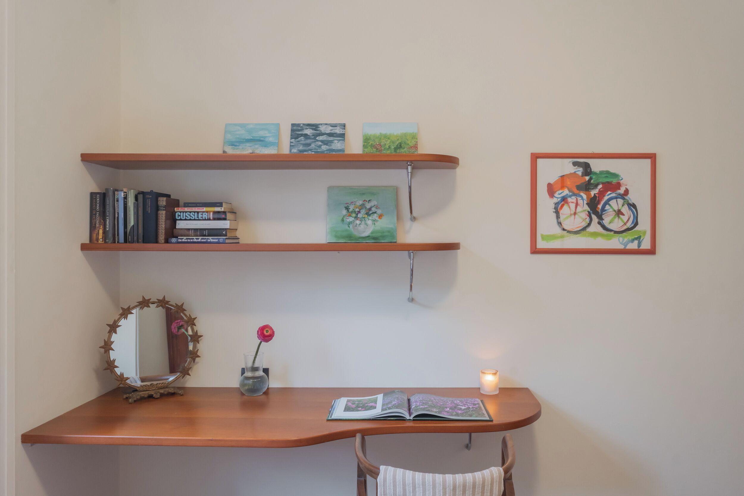 A study area with a wooden desk, books, a mirror, a vase with a flower, and artwork on the wall.