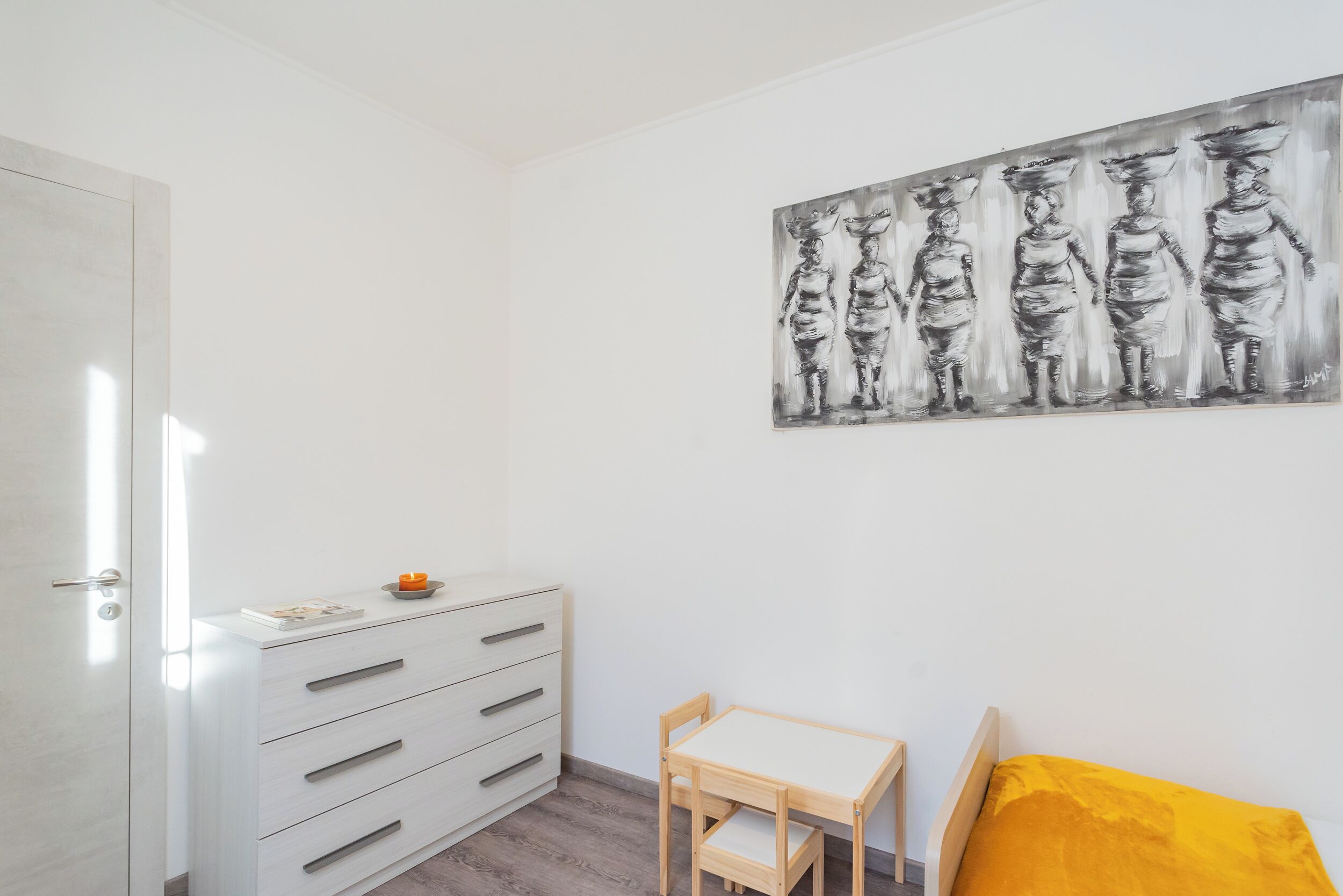 A room with a white dresser, a small wooden table, a yellow pillow, and a black and white painting on the wall.