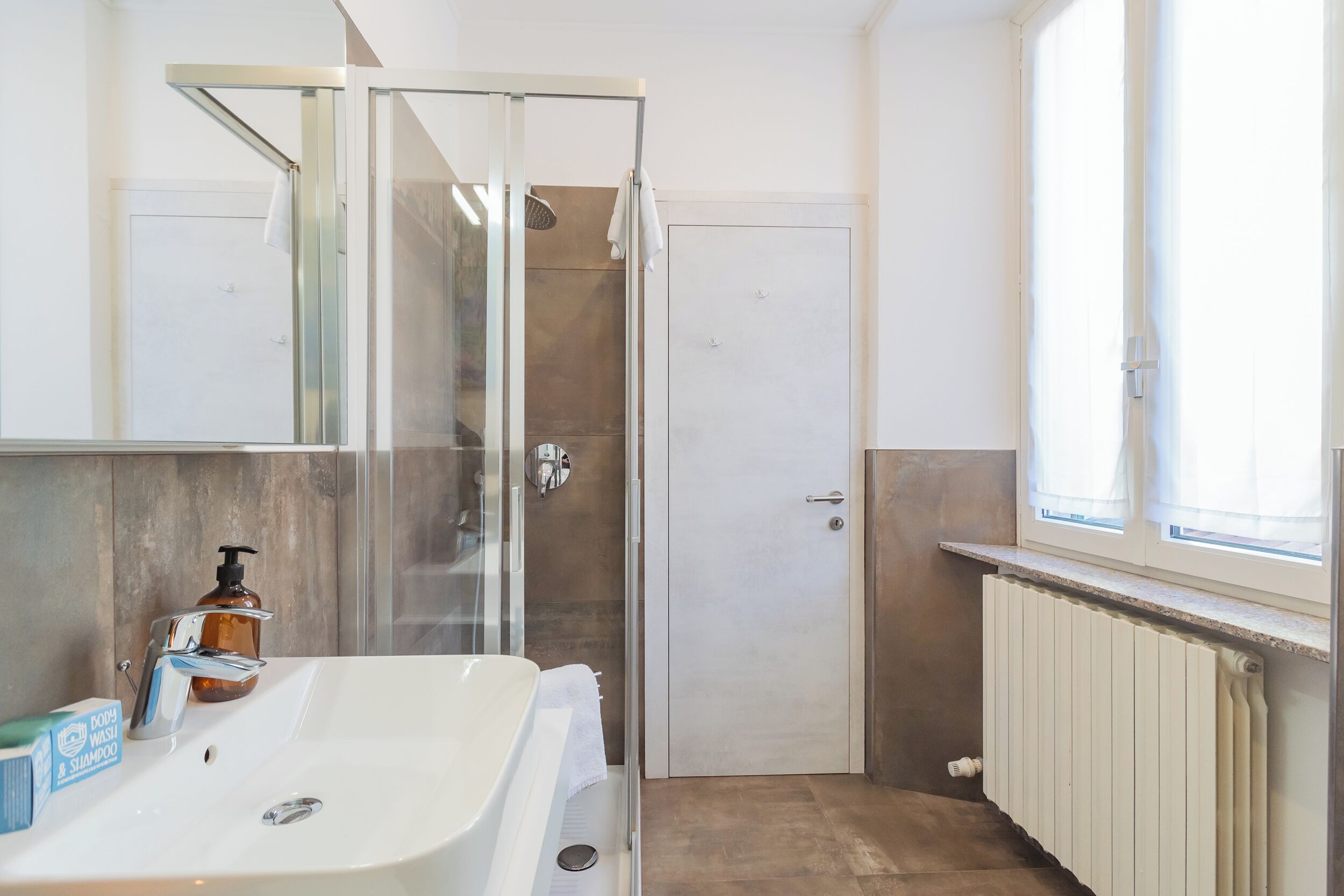 A bathroom with a glass-enclosed shower, a white sink with a soap dispenser, and a window providing natural light.