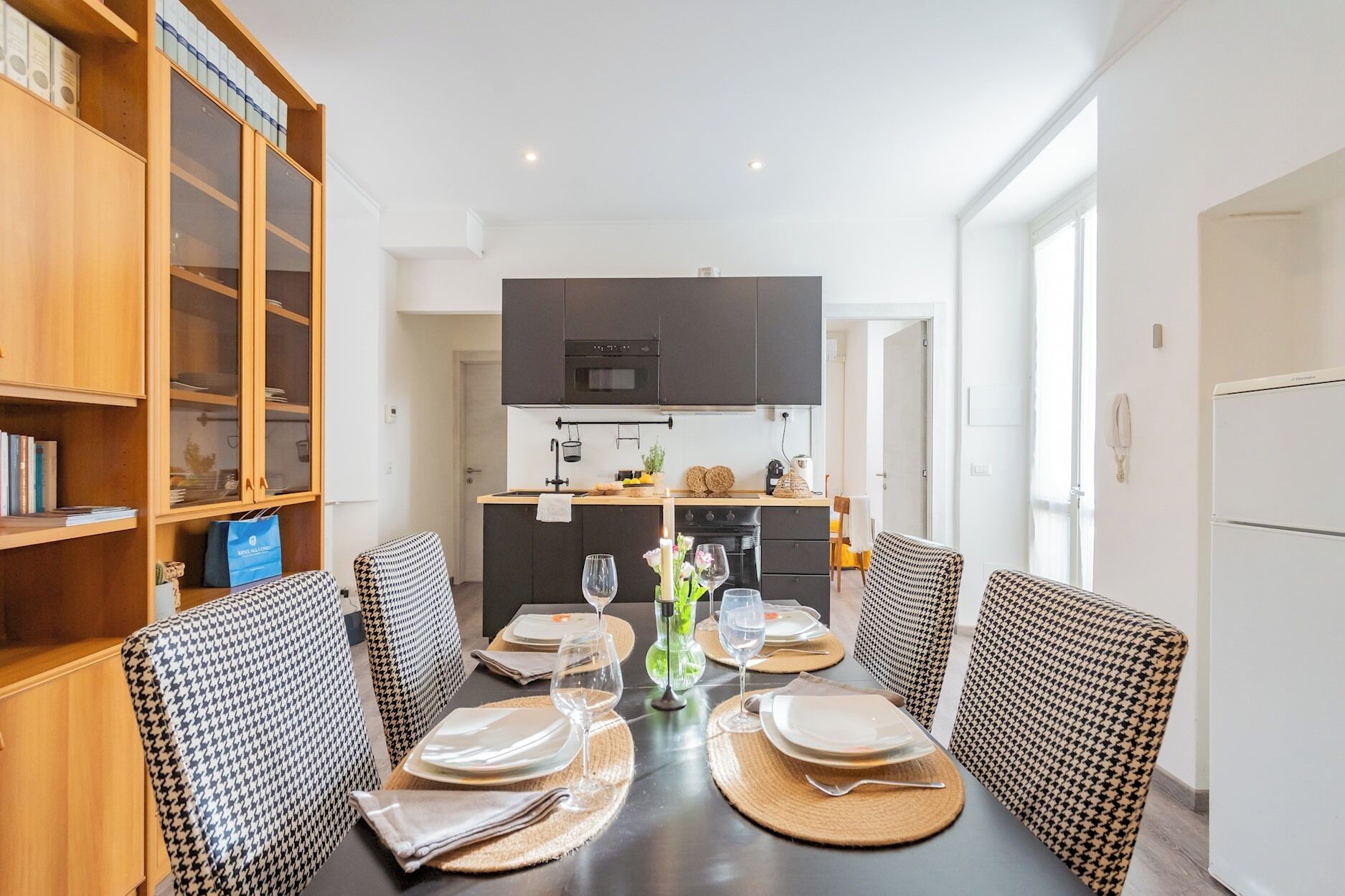 A dining area with a dark wooden table set for four, surrounded by houndstooth patterned chairs. A kitchen with dark cabinets and a microwave is visible in the background.