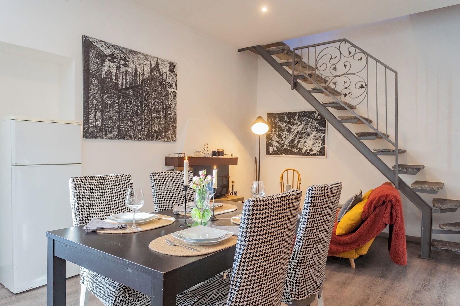 Un comedor con una mesa dispuesta para cuatro, sillas a cuadros de galgo, una mesa negra y una escalera con barandilla de metal.
