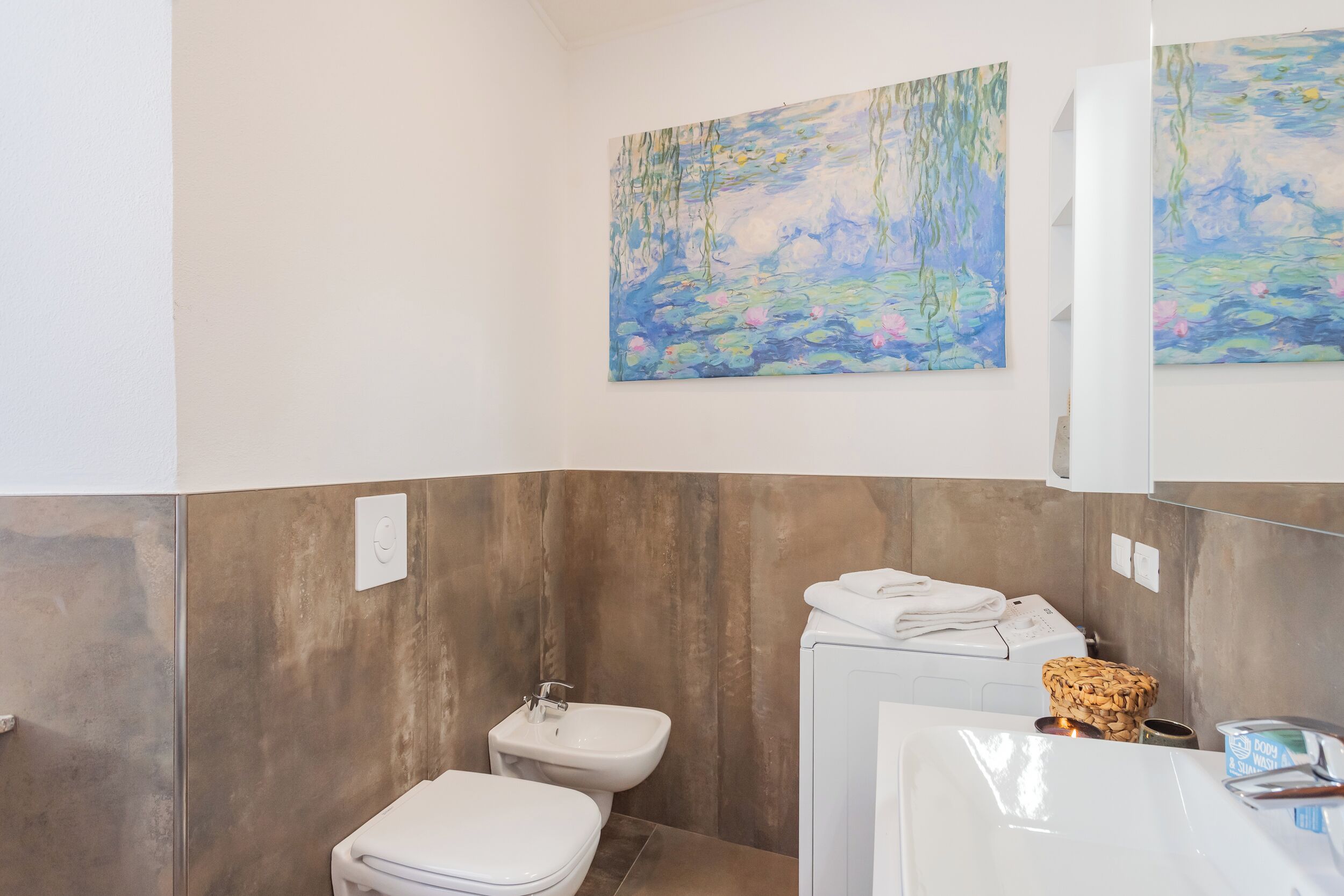 A modern bathroom with a wooden panel wall, a white sink, a toilet, and a bathtub. There are two paintings on the wall above the bathtub.