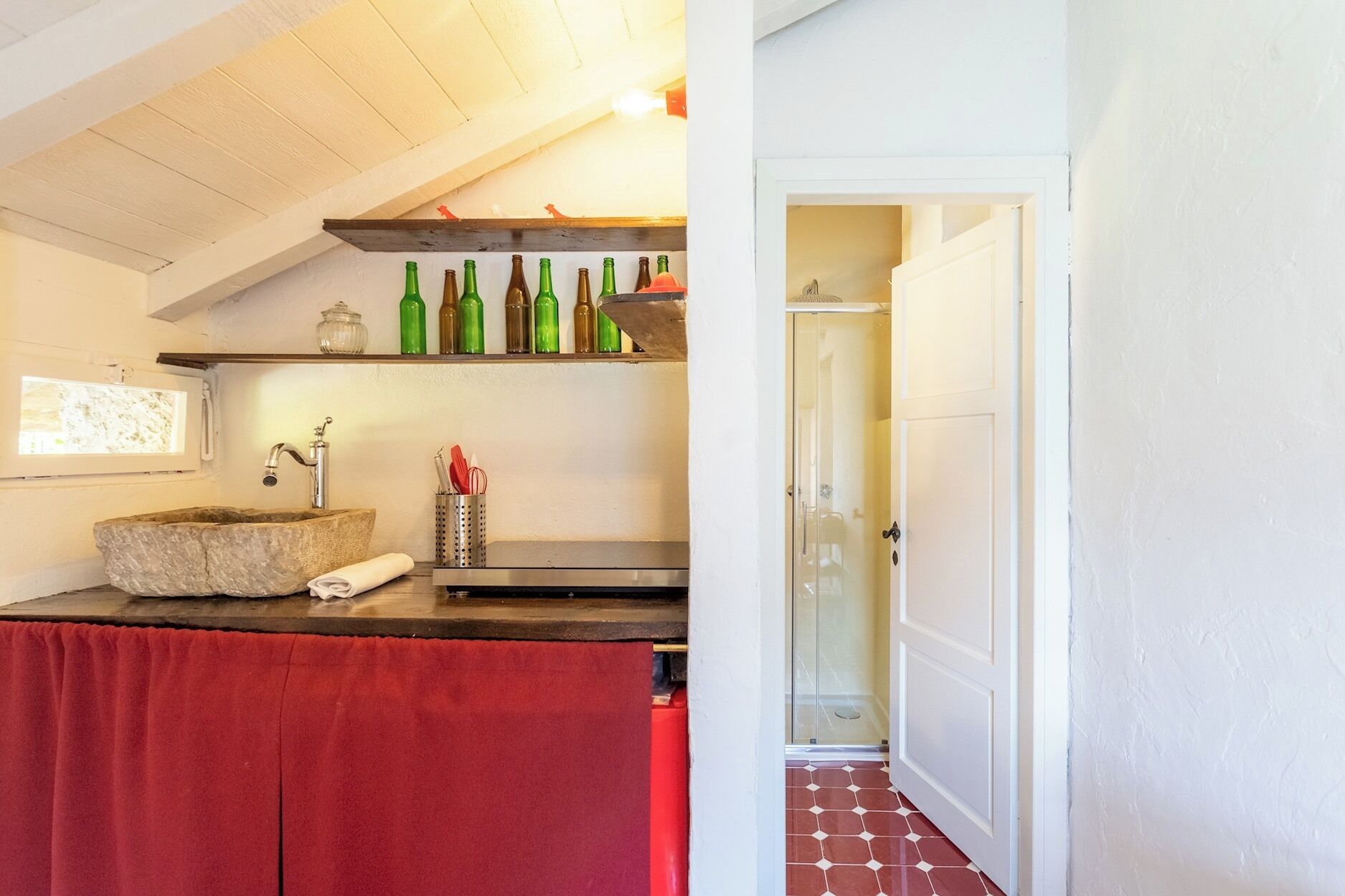 A bathroom with a stone sink, a red towel, a shelf with bottles, and a shower.