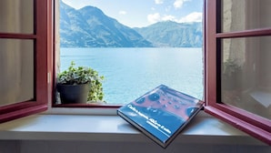 A window with a view of a lake and mountains, a plant on the windowsill, and a book titled "Cercetto in Liguria, Riva il mito".