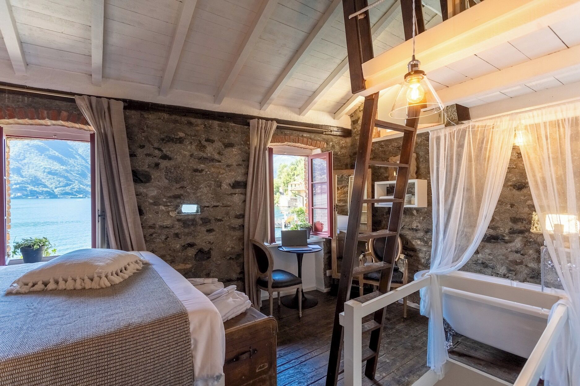 A rustic room with a bed, a desk, a chair, a ladder, and a bathtub.