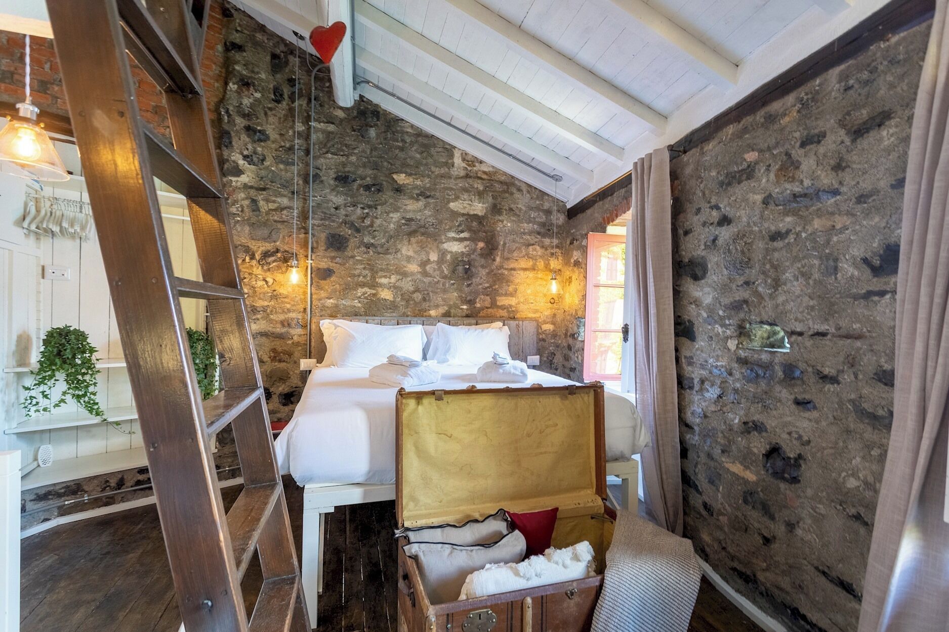 A rustic room with a stone wall, a bed with white linens, a wooden ladder, and a suitcase.
