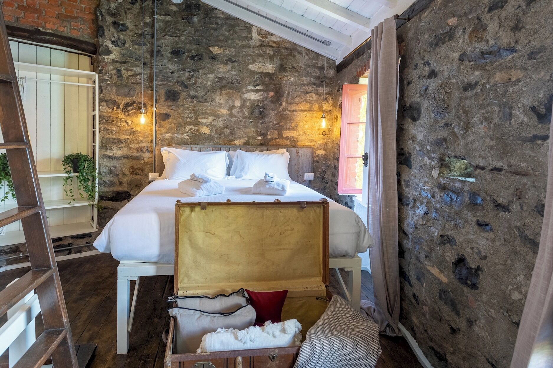 Une pièce rustique avec un lit, une échelle, une valise et un mur en pierre.