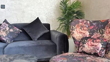 Un salón con un sofá gris oscuro, dos cojines estampados con motivos florales, un sillón a juego y una mesita redonda pequeña.