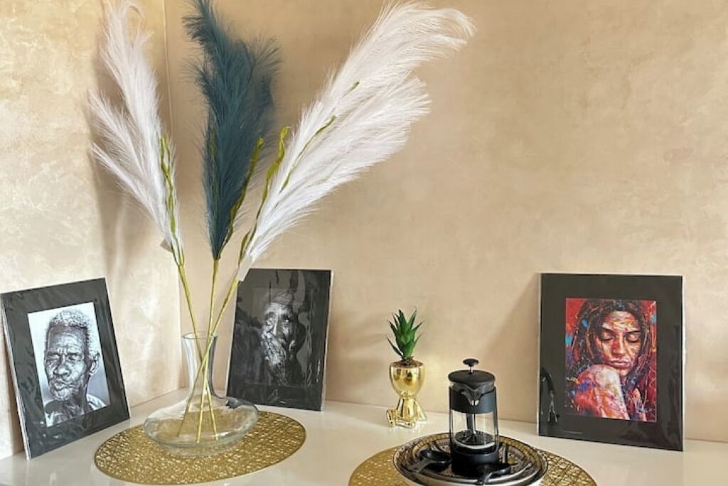 Une table ornée d’un vase de plumes, d’une petite plante et d’une machine à café, le tout entouré d’œuvres d’art encadrées.