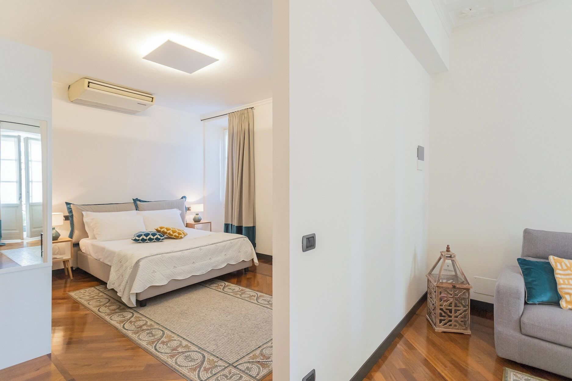 A modern bedroom with a bed, bedside tables, a nightstand, a lamp, a window with curtains, a wooden floor, and a small wooden lantern.